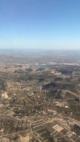 The video shows an aerial view of a landscape under a clear blue sky. A body of water, likely a lake, is visible in the distance amidst a spread-out town. The foreground shows a terrain with many fields, vegetation, and scattered houses. Roads or paths are also visible, running through the area.