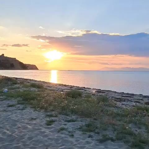 A serene beach scene unfolds at sunset. The sun, a bright orb, casts a golden reflection across the calm water, painting the sky with hues of orange and blue. Gentle waves lap at the sandy shore, bordered by sparse vegetation. In the distance, a rugged coastline rises from the sea. Two figures, dressed in white, stand near the water's edge, their poses suggesting contemplation or a ritual. As the scene transitions, two women are shown relaxing on the beach. One, with blonde hair, wears a floral dress and looks up towards the sky, while the other, with lighter hair, is draped in white and looks towards the viewer. The ambient light suggests the close of day, creating a peaceful and tranquil atmosphere.