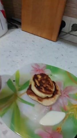 The video shows a close-up view of a breakfast scene. In the foreground, a plate with a floral pattern holds three golden-brown pancakes stacked on top of each other, accompanied by a dollop of cream or yogurt. To the right of the plate, a red mug is visible. A hand is shown dipping a tea bag into the red liquid in the mug, suggesting that tea is being prepared. In the background, a white electric kettle with a red floral design and a wooden surface are partially visible, hinting at a kitchen setting. The lighting suggests it is daytime. The overall mood is calm and domestic, focusing on the preparation and presentation of a simple breakfast. No visible text is present in the video.