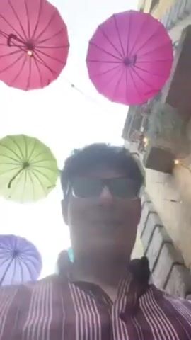 A man wearing glasses and a vertically striped shirt is taking a selfie with an upward angle. Above him are multiple brightly colored umbrellas: pink, purple, teal, and green. The background includes a building with windows and some greenery. The man is smiling slightly.