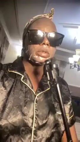 A person is standing in front of a microphone stand wearing black sunglasses, a black and gold patterned pajama top, and a patterned head wrap. They have a white substance on their face around their mouth. The background shows a chandelier, a glimpse of a sofa, and a curtained window. The person is speaking into the microphone, and there is slight movement and adjustment of the head wrap.