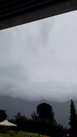 This media file captures a wide shot of a mountainous landscape under a cloudy sky. The scene is dominated by muted, cool tones, suggesting a cool or overcast day, possibly with rain imminent or having recently passed. Dense foliage, including silhouetted trees of varying heights, forms a dark band across the lower portion of the frame. To the left, a white tent is partially visible, hinting at a temporary setup like a market stall or event area. In the foreground, the ground appears to be grassy or unpaved, with some scattered orange objects visible, possibly safety cones or markers.

The backdrop is a dramatic mountain range, with its peaks shrouded in mist and heavy clouds. The darkness of the trees and the muted light create a somber and somewhat dramatic atmosphere. The overall impression is one of a natural, perhaps slightly wild or secluded location, possibly a park or a rural area at the foot of the mountains. The time of day is ambiguous due to the overcast conditions, but it appears to be daytime. There is no discernible activity of people in the frame, making the scene feel still and serene, or perhaps melancholic due to the weather. The location cues, combined with the visual elements, evoke a sense of being in a remote, mountainous region where the weather is a significant factor in the overall ambiance. The image is devoid of any visible text.