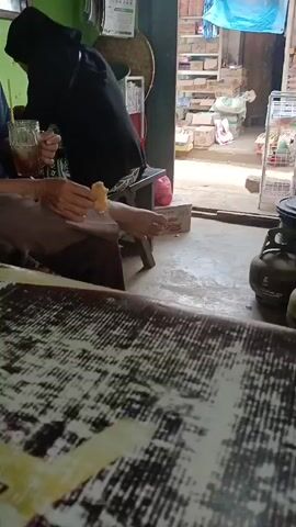 A low-angle, slightly shaky video captures a casual outdoor scene. In the foreground, the textured surface of a table, possibly made of wood with a distressed finish, dominates the lower half of the frame. The middle ground shows two people seated. The person on the left, wearing a dark headscarf and black garment, is holding a glass with a straw. The person on the right, wearing a blue shirt and brown pants, is holding a small, light-colored object, possibly food, to their mouth. Their left hand is also visible. To the right of the seated individuals, a metal gas tank is visible, along with another similar tank in green. The background reveals shelves filled with various items, suggesting a shop or market stall. A blue tarp hangs overhead. The overall setting appears to be an outdoor establishment with limited lighting.