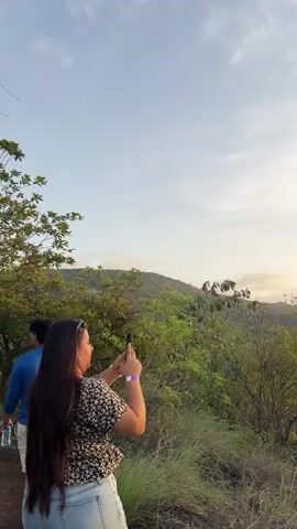 The video opens with a woman standing in the foreground, holding a phone to take a picture. She is wearing a floral top and jeans. The background features lush green vegetation and a hazy sky. As the video progresses, the camera pans to the right, revealing a sunset. The sun is a bright orange orb, partially obscured by the trees, casting a warm glow over the landscape. The sky transitions from a light blue to a soft yellow and orange hue as the sun dips below the horizon. The vegetation continues to dominate the foreground, with the silhouette of distant hills visible in the background. The video concludes with a wider shot of the sunset over the treeline, showcasing the natural beauty of the environment.