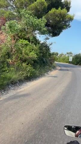 A first-person perspective video shows a motorcycle riding along a paved road that curves through a landscape of green hills and trees. The sky is blue with scattered clouds. The road appears to be in good condition. The motorcycle is moving at a moderate speed, and the rider's gloved hand is visible on the handlebars. A side mirror is also visible, reflecting a partial view of the rider and the surroundings. The scene suggests a scenic drive through a rural or coastal area.