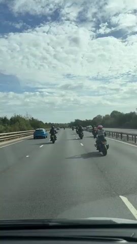A group of motorcycles are riding in formation on a highway. The motorcycles are in the right lane, and there are cars in the left lane. The sky is cloudy, and the sun is shining. The road is paved, and there are trees and vegetation on either side of the highway.