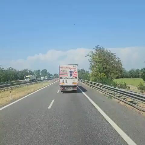 The video shows a view from a vehicle on a highway on a sunny day. The primary focus is a large white truck with advertising on its rear doors. The advertisement features the text "WERDE MIT UNS ZUM LOGISTIKHELDEN" which translates to "BECOME A LOGISTICS HERO WITH US." Below this, it states "WIR SUCHEN AUSZUBILDENDE" meaning "WE ARE LOOKING FOR APPRENTICES." It then lists job titles: "Speditionskaufmann/-frau" (Shipping Clerk), "Fachkräfte für Lagerlogistik" (Warehouse Logistics Specialists), and "Berufskraftfahrer/innen" (Professional Drivers). There is an illustration of a person dressed as a superhero. The truck has the company name "TERRA TRANS" displayed prominently. There are also license plates and other markings on the back of the truck. The truck is in motion on a highway with other vehicles visible in the distance. The environment is a highway with a guardrail on the right side and trees and fields beyond. The sky is blue with some clouds.