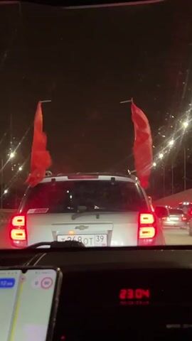 A car is driving on a road at night, with multiple street lights on. A silver Chevrolet Captiva SUV is directly in front, with two red flags sticking up from the top. The license plate of the car reads "T268OT 39". The time displayed on the car's dashboard is 23:04. A smartphone is on the dashboard, with a navigation app open. The road is congested, and other cars are visible.