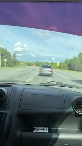 The video shows a view from inside a car, looking through the windshield at the road ahead. Initially, a gray Lada car is directly in front of the camera car. The Lada has a license plate that reads "P 754 YT 18." The cars are traveling on a paved road with double white lines. The environment consists of a rural landscape with green fields and a few trees under a partly cloudy sky. As the vehicles approach an intersection, the Lada slows down. A blue car is also visible. The gray Lada turns left. The camera car then continues forward. There are road signs and lampposts along the roadside.