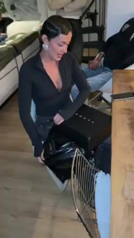 A woman sits on a white plastic chair in what appears to be an apartment with wooden floors, opening a black box labeled "GUAPE". She has short dark hair, is wearing a black long-sleeved top, jeans, and large earrings. She smiles as she pulls the box out of a black plastic bag.  Behind her, other people are visible, some seated on a bed and others standing. A wooden table is to her right.  Stairs are visible in the background.