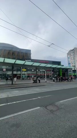 The video shows an outdoor scene, likely a public transport hub or a street in a European city. There is a bus station with several buses, and people are waiting. There are shops with signs like "TEDI", "TK Maxx", and "Media Markt". The architecture of the buildings suggests a modern design with a glass-covered walkway over the bus stops. Overhead, there are numerous power lines, indicating the presence of trams or trolleybuses. The sky is cloudy, but the sun is also visible, creating some glare. The ground is paved with asphalt and cobblestones. Several individuals are walking, standing, and some appear to be boarding or disembarking from the buses. The overall impression is of a busy urban environment.