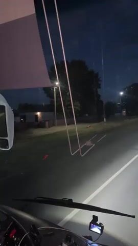 This is a first-person perspective video taken from the windshield of a vehicle, likely a bus or large van, at night. The road ahead is illuminated by the vehicle's headlights, with a white line marking the lane. In the distance, other vehicles' headlights can be seen. The sky is dark blue with some clouds and streetlights are visible along the side of the road. Trees and some buildings are dimly lit in the background. There is a small dashboard camera visible in the lower part of the frame. The scene suggests travel on a highway or main road outside of a populated area, given the presence of trees and the relatively sparse development visible. The time of day is clearly night, and the weather appears to be clear. The mood is one of quiet travel, with the focus on the road ahead.