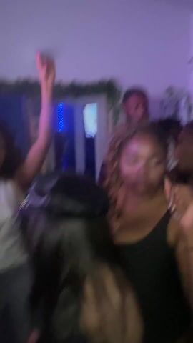 The video shows an indoor party with several people dancing in close proximity. The lighting is dark with blue and purple hues. In the foreground, a person holds a red drink in a white cup, and another person in a black lace top and a studded hat looks toward the side. A woman with curly hair wears a black tank top. In the background, a man with a beard is visible. Greenery decorates the wall. The overall atmosphere seems lively and energetic.