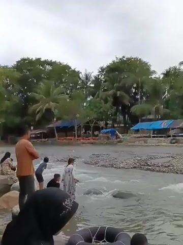 The video shows a group of people at a riverbank. In the foreground, a person in a black head covering is partially visible. To their left, a child in a blue shirt and dark shorts is playing in the water. Beside the child, a girl in a white dress with a floral pattern is also in the shallow water, appearing to be interacting with the child. Further to the right, a man in a red shirt stands in the water, looking towards the right side of the frame. The river has a rocky bed and the water is flowing. In the background, there are several structures that appear to be stalls or small buildings with thatched or corrugated roofs, set against a backdrop of lush green trees, including palm trees. The sky is overcast. The overall scene suggests a relaxed outdoor setting, possibly a riverside recreational area.
