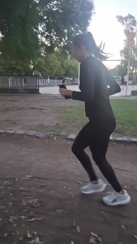 A woman in a black top and leggings runs on a dirt path in a park. She has a ponytail and wears light-colored sneakers. She's holding a cell phone in her left hand. In the background, people are playing soccer on a field. Trees line the perimeter of the park, and a fence is visible. The lighting suggests it's late afternoon or early evening.