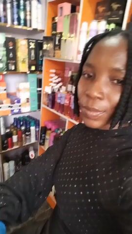 A medium shot shows a Black woman with braided hair and a black, sparkly top. She is in a store with shelves of perfumes and cosmetics behind her. She holds a few small perfume bottles up to the camera and smiles. The lighting is bright and the background is slightly blurred.