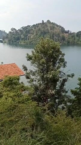 The video shows a lake surrounded by lush vegetation. Several houses are visible on the shore, with one having a distinct orange-tiled roof. The lake appears calm, and there is a floating platform in the water. The scene is taken from an elevated position, offering a panoramic view of the lake and its surroundings. The sky is overcast.