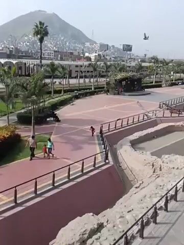 The video shows an elevated view of a park-like area, possibly in a city. In the foreground are exposed stone structures enclosed by a low wall and black metal railing. Adjacent to this is a paved walkway, also bordered by railings, that leads into a public square with a stage.  Further in the background is a long, narrow pool with several small fountains.

Several people are visible. A man and two children are walking along the path.  A boy runs across the square. Other people are scattered throughout the scene. Birds fly across the sky.

The square features a stage area, palm trees, benches, and paved pathways.  The scene contains a few objects: a cannon, and a restaurant.  A city skyline with a prominent mountain in the distance can be seen. The sky is bright and slightly overcast.  The overall impression is a public park with historical elements incorporated into its design.