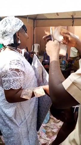 A man is handing out what appears to be Nigerian currency to two women. One of the women is wearing a white lace dress and a white head covering. The other woman is wearing a black and gold dress. The man is wearing a tan shirt. The exchange is taking place outdoors under a partially open-roofed structure. Bills of currency are already scattered on the ground.