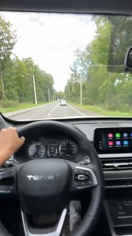 The video begins inside a moving car, showing the driver's perspective of the road ahead. The dashboard and steering wheel are visible in the foreground. The road is a two-lane highway with trees and utility poles lining both sides. Other vehicles, including cars and a white van, are visible on the road ahead. The sky is partly cloudy with patches of blue. The camera pans slightly, giving a wide view of the passing scenery.