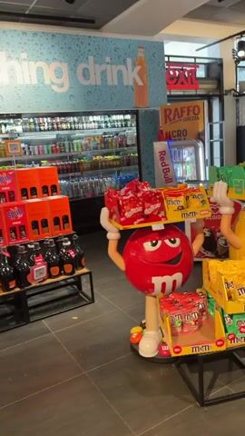 This video shows a display of M&M's candy in what appears to be a store or convenience store. Two large M&M character statues, one red and one yellow, are prominently featured. The red M&M statue is holding a shelf of M&M's bags and Maltesers. The yellow M&M statue is holding a shelf of M&M's bags and has M&M's scattered around its base. In the background, there are shelves stocked with various beverages and snacks. A sign above the beverage shelves reads "thing drink". Another sign to the right advertises a "30% SCONTO" (30% discount). A person in a blue shirt walks by in the background. The lighting suggests an indoor commercial environment.