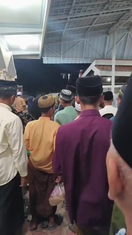 A group of people are standing in what appears to be a queue outside an indoor area. Most of the people are men wearing traditional Muslim attire, including peci hats and long shirts or robes. Some are wearing peci hats with decorative patterns. One person in the foreground is wearing a purple long-sleeved shirt, and another to their left is wearing a tan-colored shirt. There is a ceiling with visible beams above the people, and lights are illuminating the area. The background is dark, suggesting it might be evening or nighttime. There are stone-like structures on the left side of the frame. The people seem to be waiting for something or moving in a particular direction.
