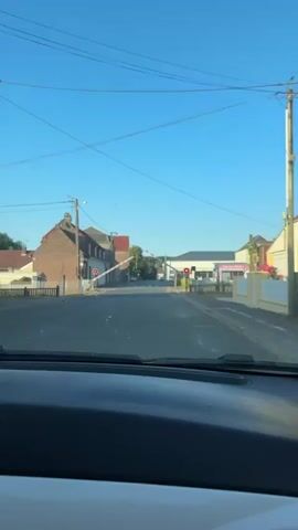 The video, filmed from inside a vehicle, shows a road passing through a village. Two railway crossing barriers are in the lowered position, indicating a train is approaching or passing. Red lights are illuminated on the barriers, signaling to stop. Buildings, including a brick building with a red roof and other residential structures, line the sides of the road. Trees are visible in the distance. The sky is clear and blue.