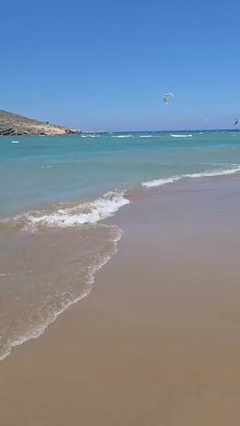 The video opens with a close-up of waves gently lapping onto a sandy beach under a clear blue sky. As the camera pans slightly, a few kite surfers are visible in the distance, their colorful kites dotting the turquoise water. The shot then moves to show a man and a woman standing near the water's edge. The man is crouched down, appearing to examine something on the wet sand, while the woman stands beside him, looking out at the sea. Several other people are scattered along the beach, some walking, others standing or sitting. More kite surfers are visible further out in the water, and a hilly, arid landscape forms the backdrop to the scene. The camera then pans to a man in patterned swim shorts walking away from the viewer on the beach, his shadow stretching out before him. Throughout the video, the sun is shining brightly, creating a warm, summery atmosphere.