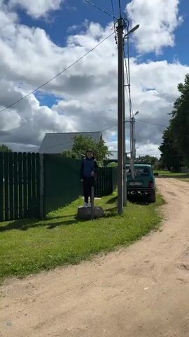 The video shows a young man standing on a concrete block next to a utility pole and a green picket fence. He is wearing a dark blue vest, dark pants, and white shoes. A young girl wearing a pink sweatshirt, jeans, and a white baseball cap walks toward him on a dirt road. She runs briefly before slowing to a walk. The background includes a house with a gray roof, green foliage, and a green car parked nearby. The camera then pans to show a dirt road with a car parked next to a wooden fence. The sky is blue with scattered clouds.