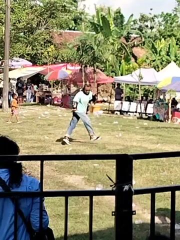 A man in a light blue t-shirt and jeans is dancing on a grassy field.  The man is moving his arms and legs in a jerky fashion. Behind him is a row of tents or stalls, some of which are shaded by colorful umbrellas.  There are other people milling around in the background. A young boy runs across the field in front of the dancing man. A person in a blue shirt stands behind a metal railing in the foreground watching. Trees and rooftops are visible in the background.
