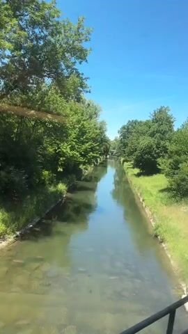 The video captures a sunny day with a clear blue sky in Glattbrugg, Switzerland. The scene begins by a tranquil canal lined with lush green trees and grass. The water is calm and reflects the surrounding foliage.

The perspective then shifts to a view from a moving vehicle, likely a train or car, as the scenery glides by. We see more greenery, including large trees and bushes. There are some objects covered with white tarps, suggesting storage or protection of items.

Further along, a concrete wall and fence come into view, bordering a grassy area. Some industrial-looking buildings are visible in the background. Graffiti adorns one of the buildings, with visible text including "USE" and "PUBER". Another structure, possibly a factory or warehouse, with a metal roof supported by pillars, is also seen. More graffiti is present on this building as well. The video ends with a view of more trees and a concrete barrier, suggesting the journey continues through an urban or semi-industrial area.

The overall impression is of a pleasant, sunny day in a mixed urban and natural landscape, with elements of industrial activity and outdoor recreation. The time of day is clearly daytime, and the weather is fair and bright.