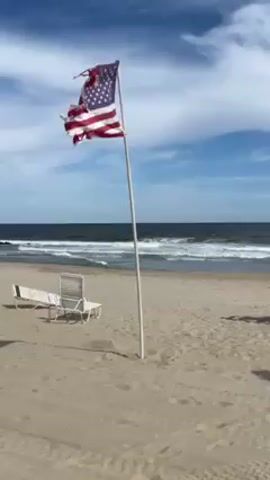 The video shows an American flag flying on a white pole on a sandy beach. The flag is tattered and torn. Two white beach chairs and a beach lounger sit on the sand behind the flag pole. The ocean is in the background with some waves and a horizon line. The sky is blue with scattered clouds.