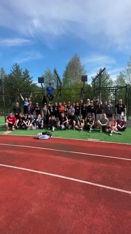 A medium shot shows a group of about 30 people taking a group photo outside on a sunny day. Most of the people are sitting or squatting on a green artificial turf surface, with a red running track in the foreground. The people are dressed in workout clothes, and some have medals around their necks. A person in a blue shirt is standing on a climbing rig in the background, behind the group. There are trees visible in the background, and the sky is blue with scattered clouds. Some of the people are raising their arms in the air as someone yells, presumably to signal for the picture.