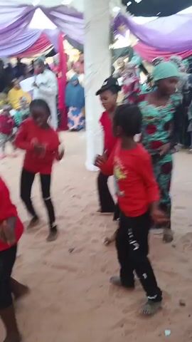 In a video captured at ground level, a group of four young Black girls are dancing in formation in a sandy area. They are dressed similarly in red long-sleeved shirts and dark pants, and most are barefoot. The camera moves to keep them centered, showing their coordinated, rhythmic steps. 

Behind them, a small crowd of adults watches. A man in a white robe and hat appears to be speaking or singing into a microphone, suggesting the dance is part of a performance. The adults are diverse in their attire, some in Western clothing and others in traditional African garments. 

The setting looks like an outdoor event, possibly a celebration. A decorated tent, draped with white and lavender fabric, provides some shade. A large white pillar is visible, further indicating a planned event. The overall atmosphere seems festive, with music playing and people enjoying the dance performance.