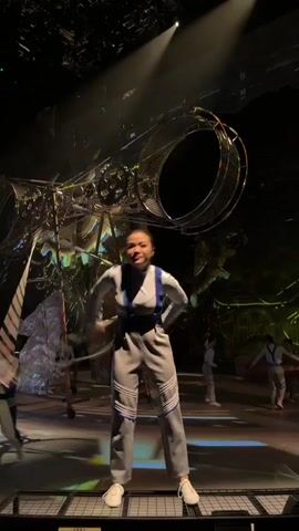 A woman in a grey jumpsuit with blue accents and white shoes stands on a stage, holding a large hoop with green and white stripes. She is in the center of the frame, and the hoop is extended above her head. In the background, there are other performers dressed in similar costumes and a large, complex metal structure that appears to be part of the stage set. The lighting is dramatic, with spotlights illuminating the central performer and creating shadows on the stage. The overall atmosphere suggests a theatrical performance or circus act.