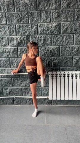 A young girl in athletic wear is practicing a dance move against a brick wall. She is balancing on one leg, with her other leg extended straight up in the air, forming a split. Her left hand is resting on a horizontal bar, which appears to be part of a dance barre or railing. To her right, a white radiator is visible. The girl is looking to her left, and her body is angled towards the left side of the frame. The lighting suggests an indoor setting.