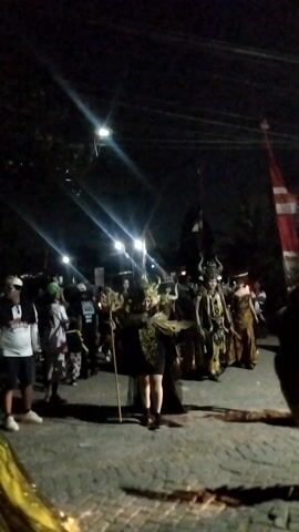 This video captures a nighttime street scene with a procession. Several individuals are dressed in elaborate, traditional costumes, some with headdresses and ornate garments, holding what appear to be banners or poles. The procession is illuminated by string lights overhead. In the foreground, a group of people in casual attire are observing the procession, some appearing to be filming or taking pictures. The ground is paved with cobblestones. The overall atmosphere suggests a cultural or ceremonial event taking place after dark.