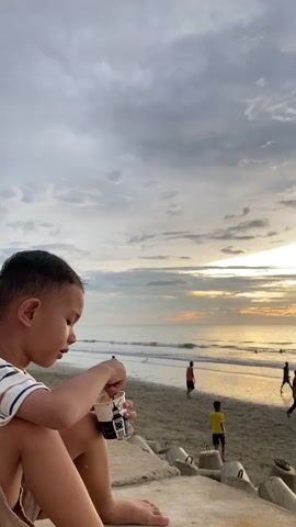 A young boy sits on a concrete structure at the beach eating from a cup. The ocean is in the background with waves breaking on the shore. Several figures, appearing to be young boys, are playing soccer on the beach. The sky is cloudy with the sun beginning to set, casting a warm glow over the scene.