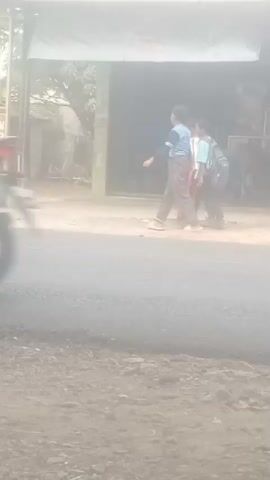A truck is visible on the left side of the frame, partially obscured by dust. The truck has an advertisement on its side featuring an image of a person. In the center of the frame, three individuals are walking across a road. They appear to be young males, and at least two of them are carrying backpacks. They are walking towards an archway that has text written on it. The archway appears to be an entrance to a compound or building. The environment is dusty, suggesting it might be a dry or unpaved area. The road in the foreground appears to be paved.