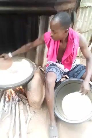 A man wearing a pink towel around his neck and patterned shorts is sitting on the ground cooking outdoors. He is stirring a large pan of white liquid over a wood fire. Next to him is another bucket containing the same white liquid. He transfers the liquid from the pan to the bucket using a wooden implement. In the background, two people are standing nearby on a dirt path. The cooking area is adjacent to a dilapidated structure.