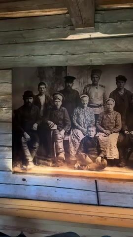 The video shows a large framed photograph of a group of people posing for a portrait. The photograph is sepia-toned and appears to be old, with a vintage aesthetic. The people in the photograph are arranged in two rows. The front row has a young boy sitting cross-legged. Behind him, on a raised platform, are several adults, including men and women, dressed in traditional clothing. The background of the photograph is a painted studio backdrop, possibly depicting trees or foliage. The photograph is mounted on a wooden wall, with visible wooden beams and planks above and around it. The lighting in the video suggests an indoor setting, possibly a museum or a historical exhibit, as the photograph is illuminated and displayed prominently.