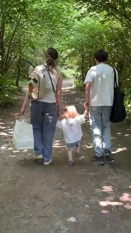 A family walks down a wooded path. The parents, dressed in jeans and white t-shirts, hold the hands of their toddler, who is wearing a white shirt, blue and white striped shorts, and metallic brown shoes with pink socks. The mother carries a white shopping bag and a beige backpack. The father carries a black bag over his shoulder. The path is dirt and gravel, and the sun filters through the green trees overhead. The camera focuses on the toddler's feet and the ground as they walk.