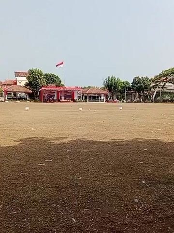 The video shows an outdoor field with a dirt and dry grass surface. In the background, there are several buildings, trees, and a flag pole with a flag. There are also some tents and carts set up, suggesting a market or event. A few people are visible, one standing near a tent and another near some buildings. The overall atmosphere appears to be festive and celebratory, possibly for a national holiday like Indonesian Independence Day (August 17th). The sky is overcast.