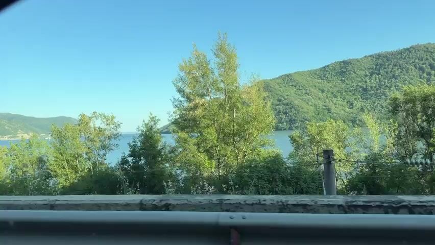 The camera looks out a vehicle window at a wide view of a blue lake or river, surrounded by green mountains and trees. The vehicle appears to be moving, as the foreground of the road and the closer trees and mountains move quickly through the frame. The sky is bright blue, indicating good weather. There are no people visible.
