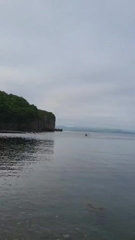A serene video captures a tranquil ocean scene under an overcast sky. The water is calm, reflecting the muted tones of the clouds. In the distance, a small boat with at least one person can be seen navigating the expanse of water. To the left, a lush, green cliff juts out into the sea, adding a natural element to the landscape. Further in the distance, several islands or peninsulas are visible, their forms softened by the hazy atmosphere. The overall mood is peaceful and contemplative. The time of day appears to be daytime, likely morning or late afternoon, given the diffused light. The weather is cloudy, suggesting a cool or temperate climate. The presence of the boat and the natural landscape hint at a coastal area. There is no visible text in the video.