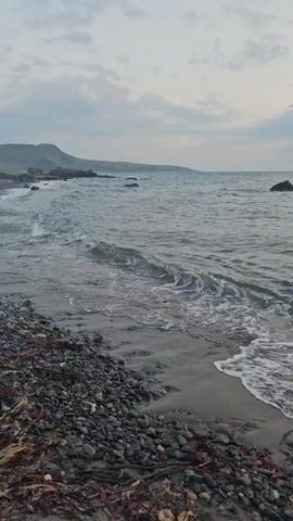 The video opens with a view of a rocky beach and the ocean. The waves gently lap at the shore. As the video progresses, the camera pans to show a bonfire on the beach. In the background, there are large rocks and the ocean. The sky is cloudy and overcast. A table and a tent are visible on the beach in the distance.