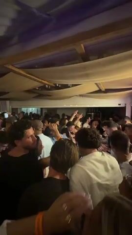 The video shows a crowded indoor party scene. Many people are packed together, facing towards the camera and behind it. Some people are holding up drinks. The lighting shifts intermittently. The ceiling is decorated with a white canopy-like structure, and there is some wooden framework visible. The overall atmosphere appears lively.