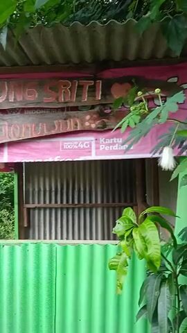 The camera pans from what appears to be a rustic building made of corrugated metal and wood. There's an overhead sign that reads "KEDUNG SRITI," and the lower section of a pink advertisement. The camera pans to reveal a view of terraced agricultural fields on a hillside. The weather is overcast or foggy. The foreground features dense foliage.