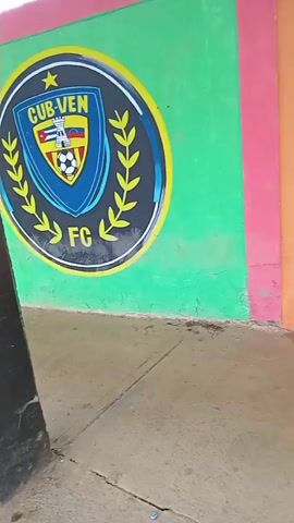 The video pans across a brightly colored mural on a wall. The mural appears to be sports-themed, with several soccer club logos visible, including "HAVEN FC", "Dinamo", and "Villanueva FC". The wall is painted in sections of green, orange, pink, and yellow. Beyond the wall, a concrete walkway leads to a covered seating area with black support pillars. The seating area overlooks a sports field, possibly a soccer field, surrounded by a fence with colorful panels. The field itself is green, and there are trees and hills in the background under a cloudy sky. The overall impression is of a somewhat weathered but vibrant community sports facility. There are no people visible in the video.