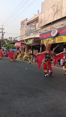A group of performers in elaborate red and gold costumes are participating in a parade on a paved street. The performers are dressed in traditional attire, with some wearing headdresses and carrying long, thin poles with leafy branches. They are moving in a synchronized fashion, some holding large red fabric pieces that resemble wings or capes. The parade is taking place during the day, with overhead power lines visible and buildings lining the street. Spectators are seated on the side of the road, watching the procession. The atmosphere appears festive.